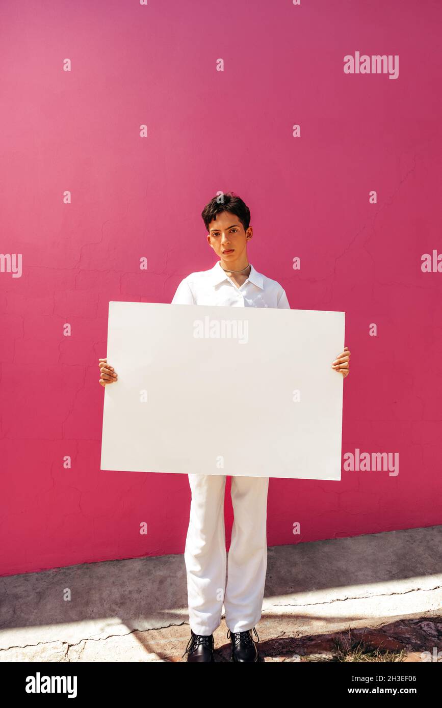 Assertive queer boy holding a white placard against a pink background. Confident teenage boy displaying a blank banner in a studio. Young gay boy comi Stock Photo