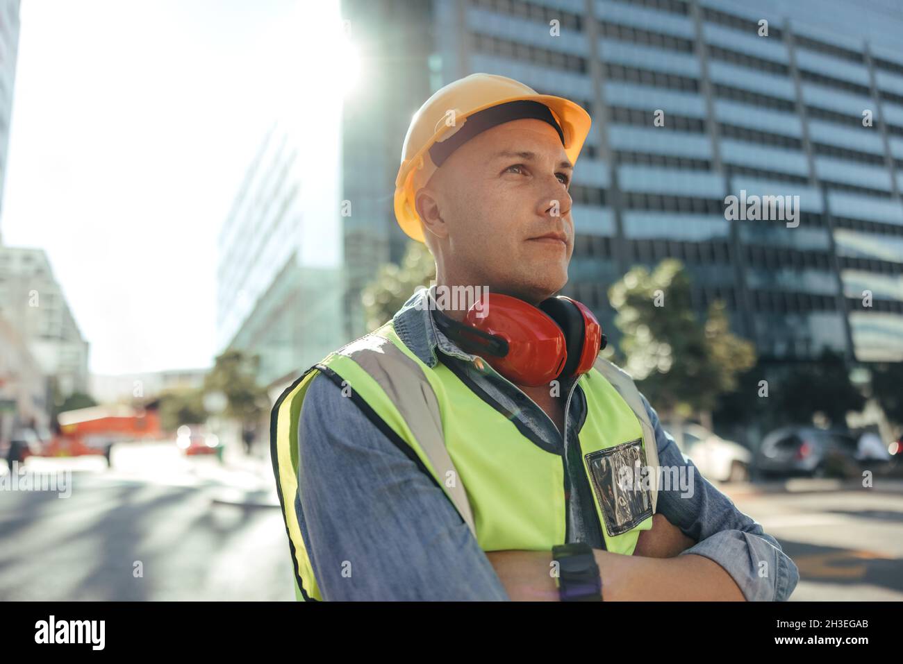 Construction worker looking away thoughtfully. Mid-adult workman standing with his arms crossed in the city. Blue collar worker standing in front of h Stock Photo