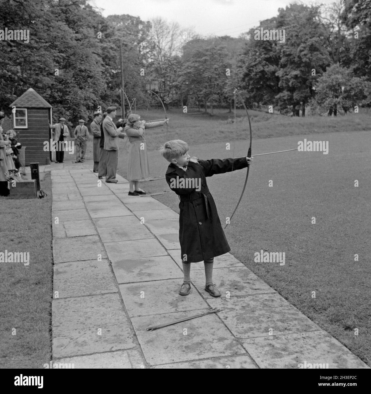A boy is amongst several people taking aim at an archery range in a park in the UK in the 1950s. He has just released the arrow. This is taken from an amateur photographer’s black and white negative – a vintage 1950s photograph. Stock Photo