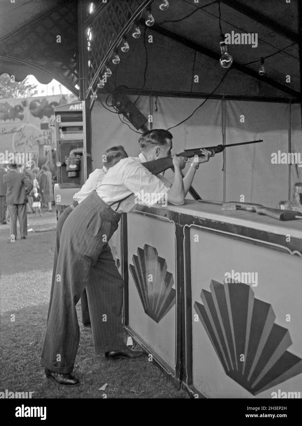 A rifle range or shooting gallery at a funfair in the UK c.1950 – here a man takes aim at a target with an air rifle. A good score or bullseye would earn a prize. Note the fairground art in the form of the ‘shell’ decorative motifs on the booth. This is taken from an amateur photographer’s black and white negative – a vintage 1950s photograph. Stock Photo