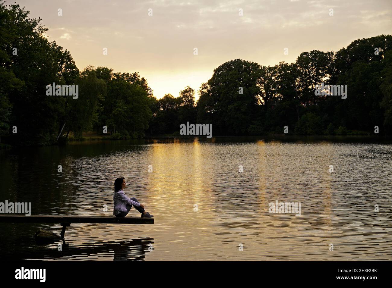 Woman sitting on a jetty by the lake. Sunset in the evening with reflection in the water. Rest and relaxation in nature. Stock Photo