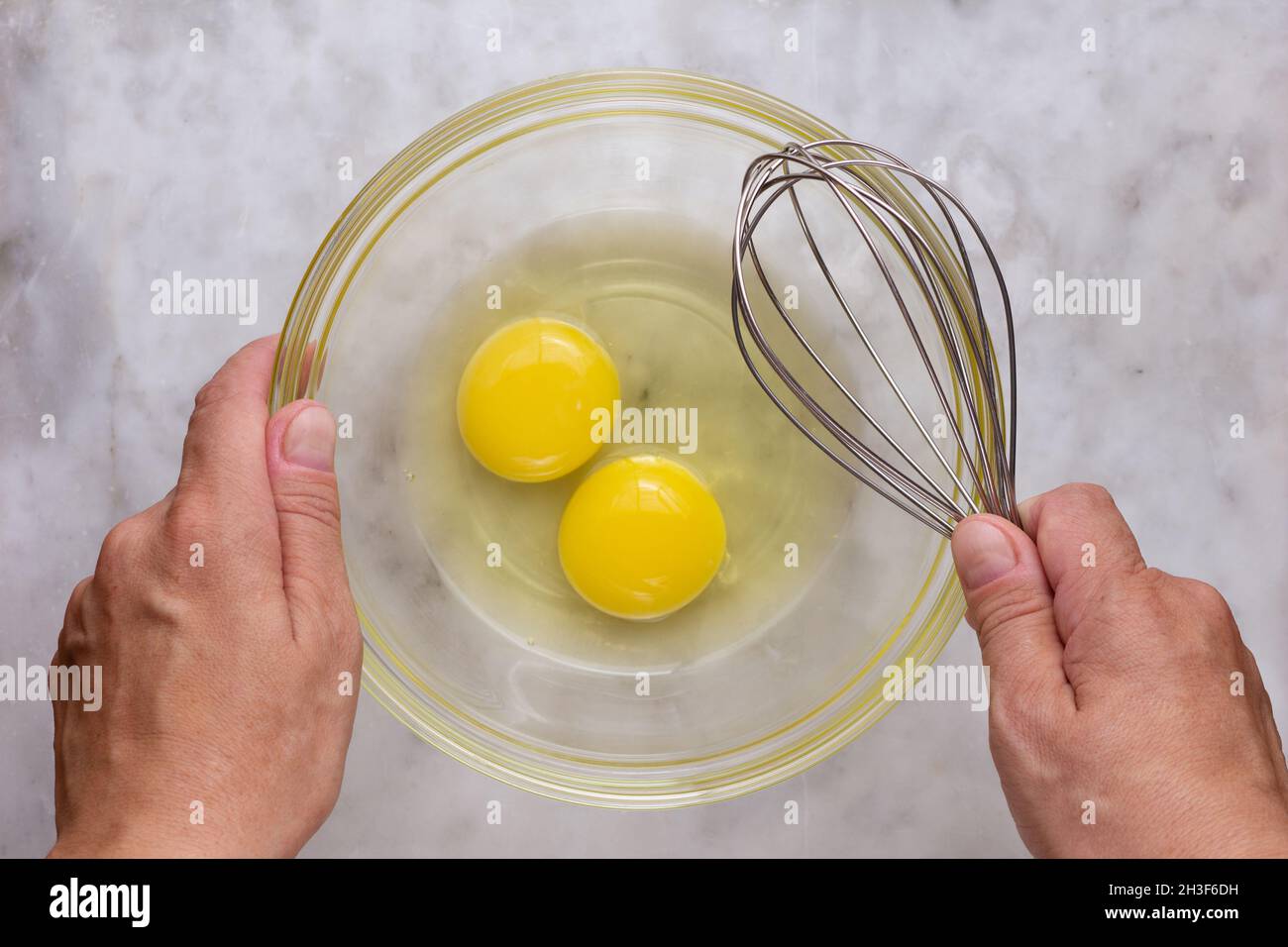 Top view of woman hands holding glass bowl with two raw yolks and whites in it and whisk on the marble surface Stock Photo