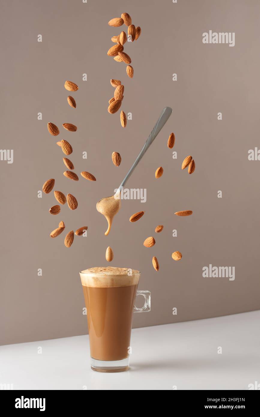 Glass cup with cappuccino and fragrant foam and a spoon and falling almonds. Cup with cappuccino, latte or coffee with milk. Creative design for cafes Stock Photo
