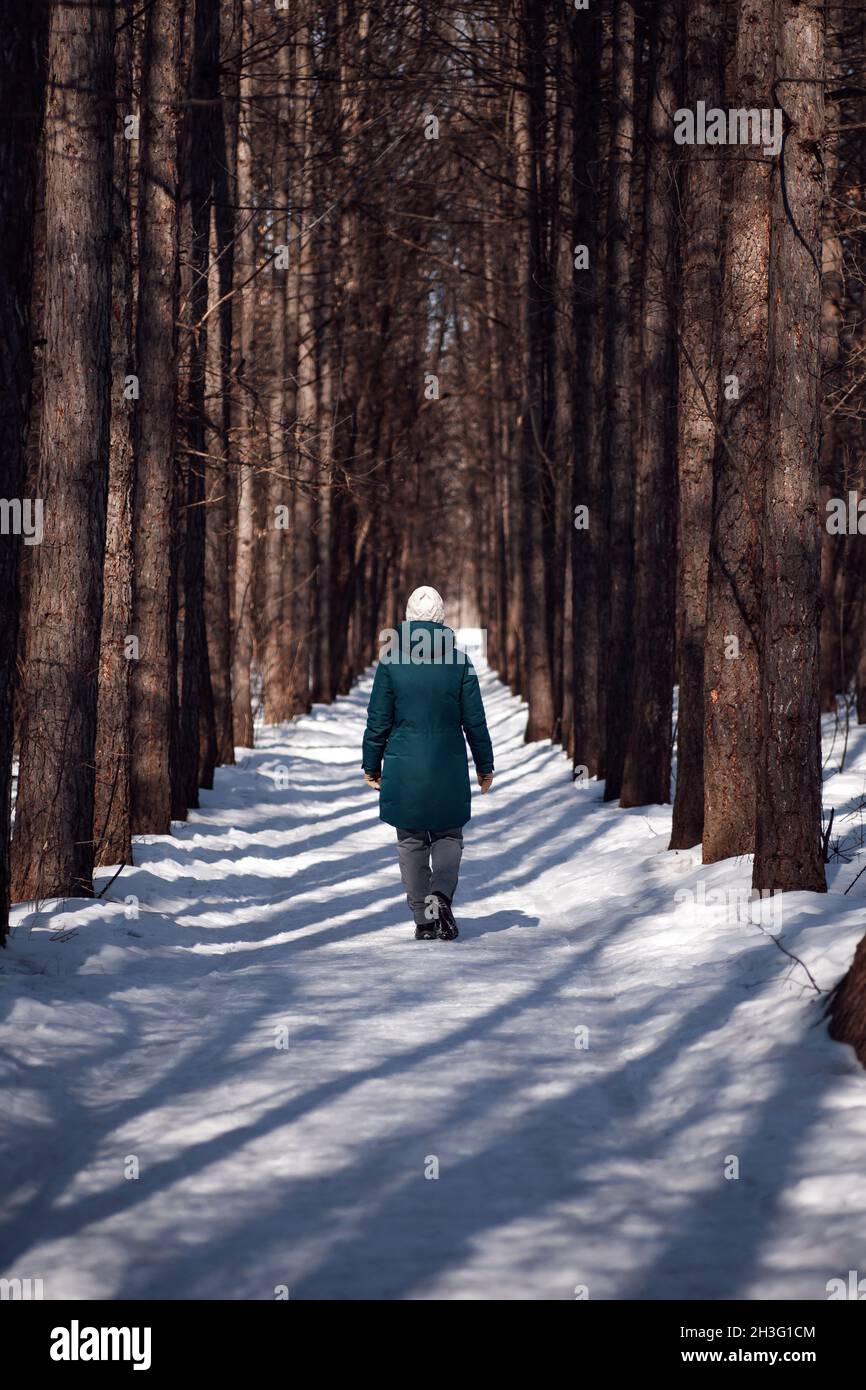 Young woman is walking along road. Winter road in forest covered with snow, young woman in green down jacket walks on beautiful frosty sunny day Stock Photo