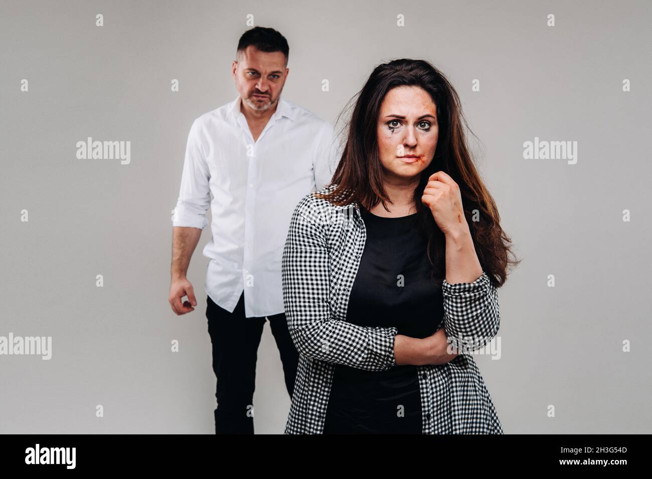 A woman beaten by her husband standing behind her and looking at her aggressively. Domestic violence. Stock Photo