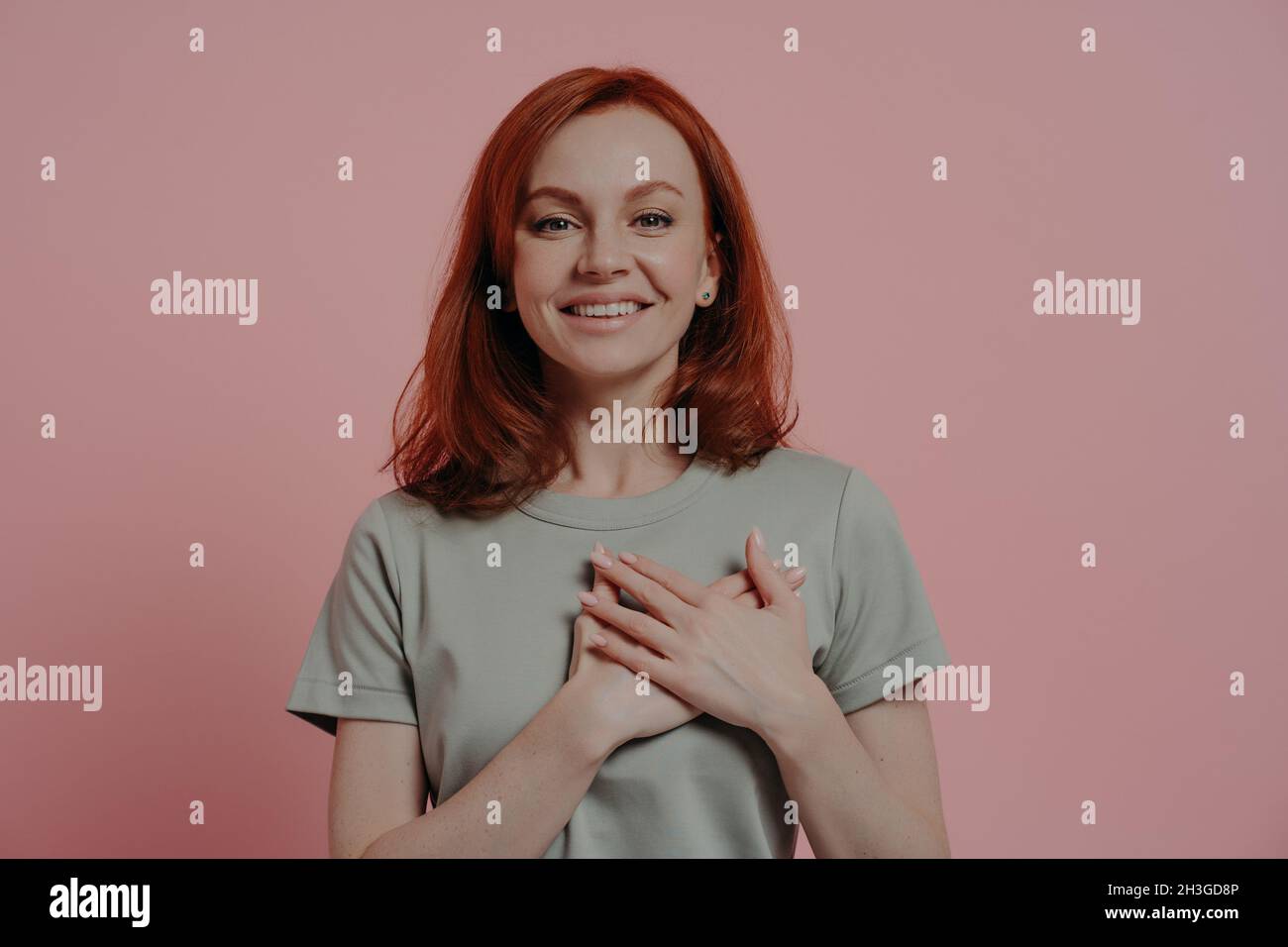 Kind lovely ginger woman keeping palms near hear and expressing thankfulness on pink background Stock Photo