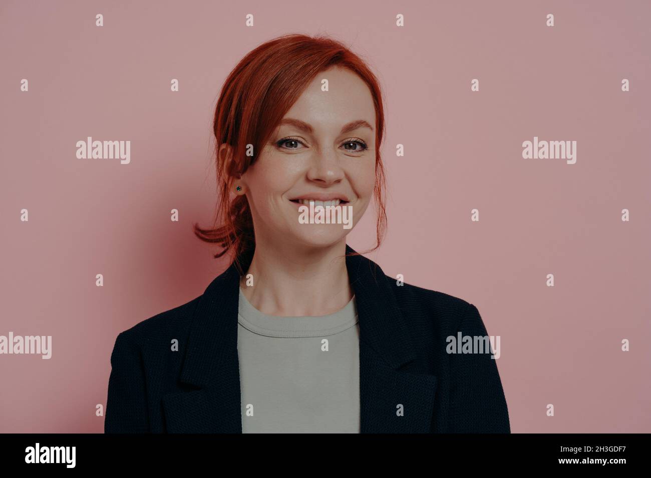 Charming young red-haired female smiling pleasantly at camera, isolated on rosy background Stock Photo