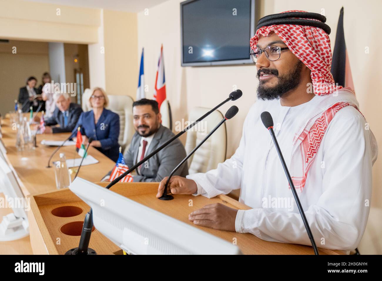 Smiling Muslim politician in ghutrah and agal standing at rostrum podium and speaking in public using mucrophone at international conference Stock Photo
