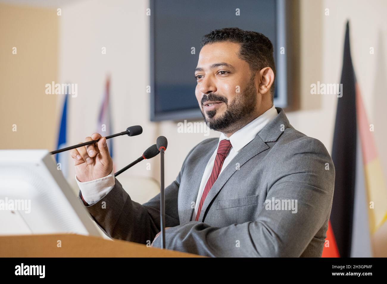 Content confident Middle Eastern politician in gray suit standing at tribune podium and speaking into microphone while presenting report at conference Stock Photo
