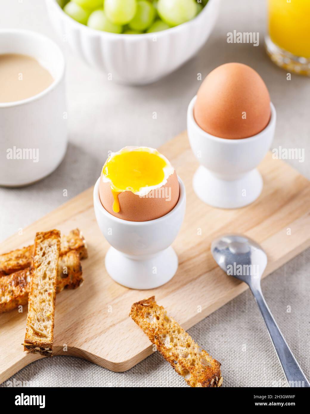 Two soft-boiled eggs in egg cups served on wooden cutting board with toasted bread for breakfast. Food background. Stock Photo