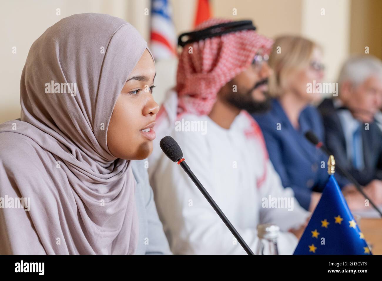 Confident young Muslim female politician in hijab speaking into microphone while nswering question of journalist Stock Photo