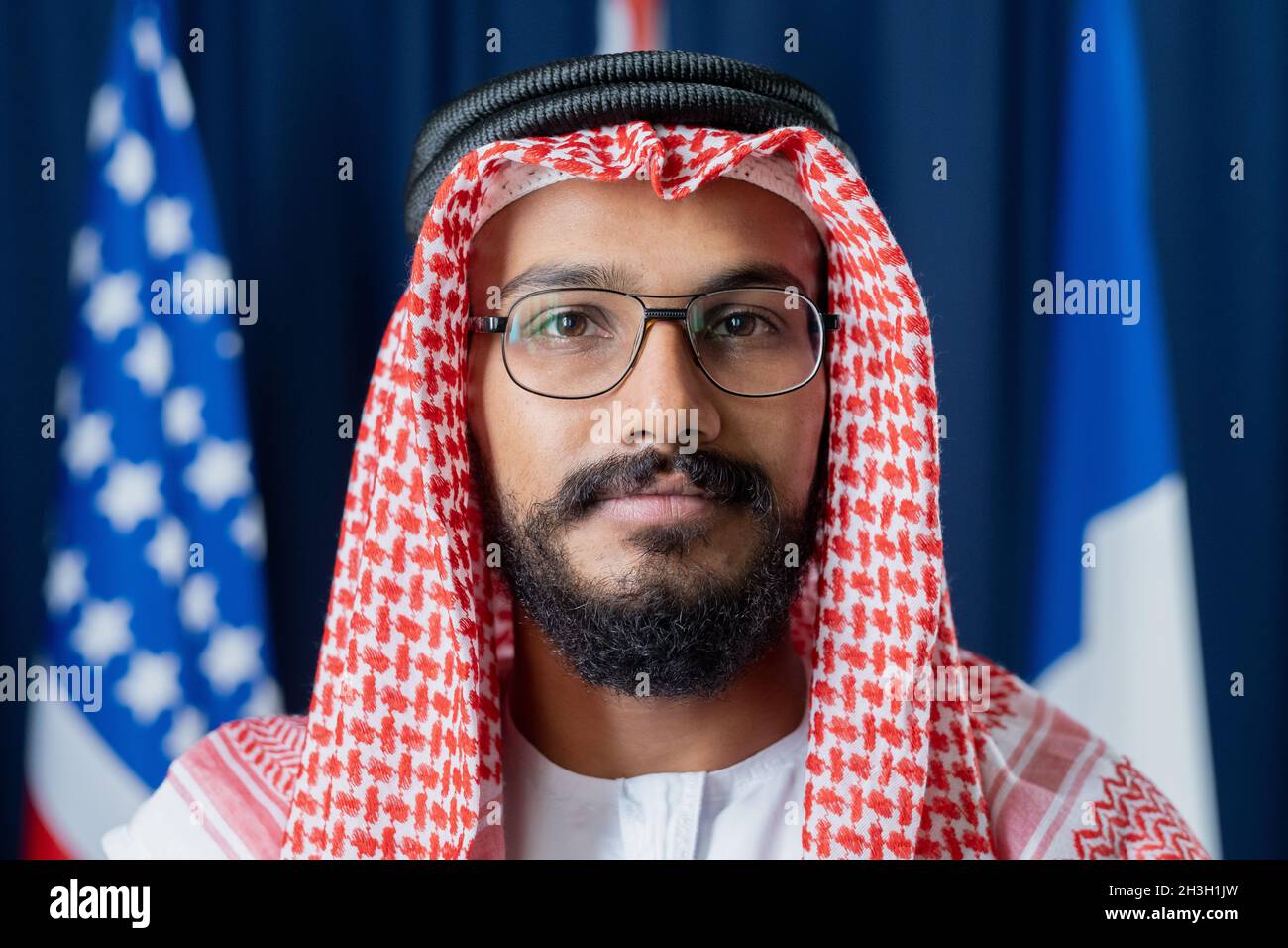 Portrait of serious confident Arabian politician in red and white ghutrah wearing agal and eyeglasses against national flags Stock Photo