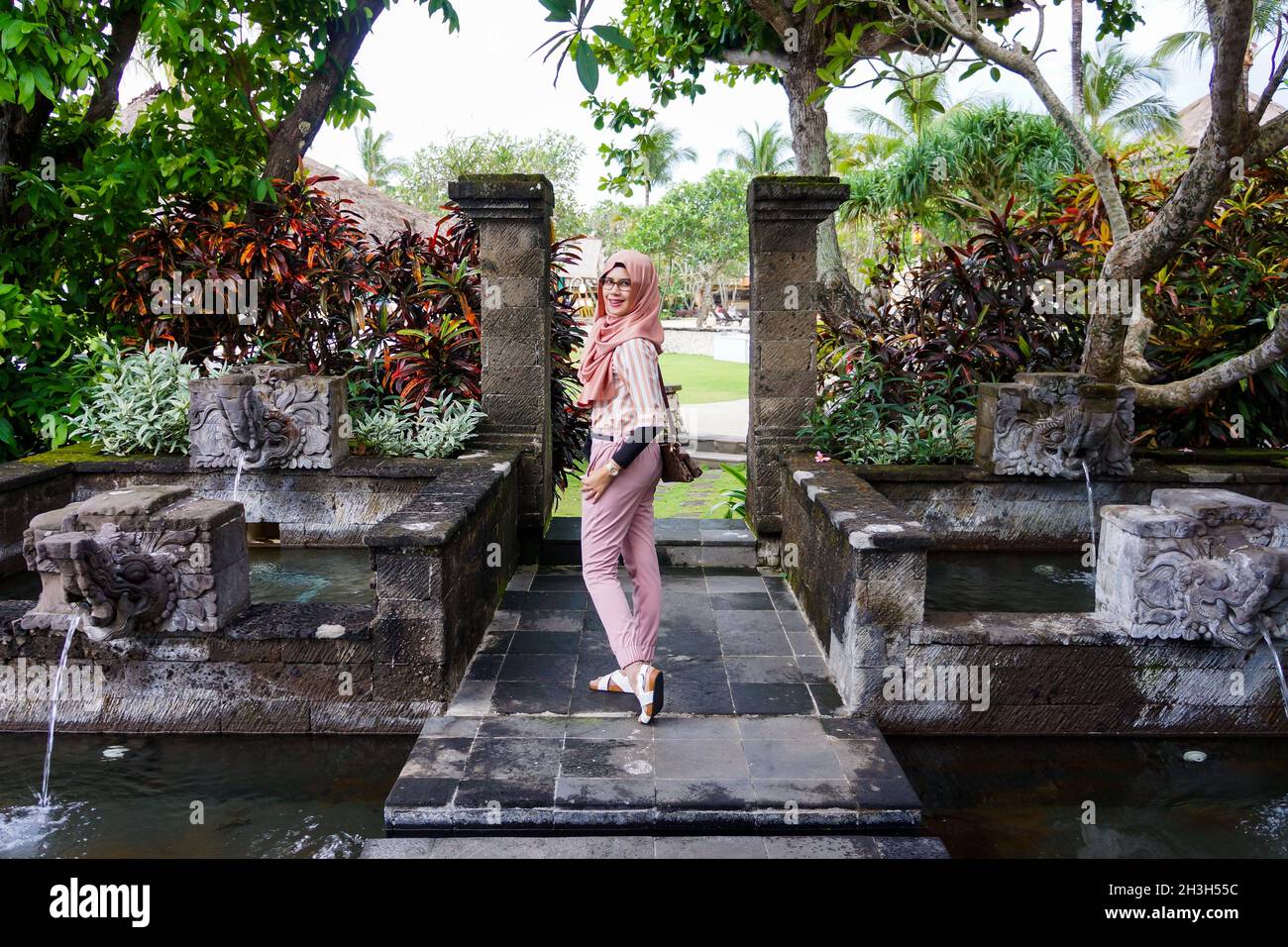 Portrait of beautiful young Asian muslim woman wearing hijab pose in garden with pond and water fountain in Bali. Smiling and happy expression. Stock Photo