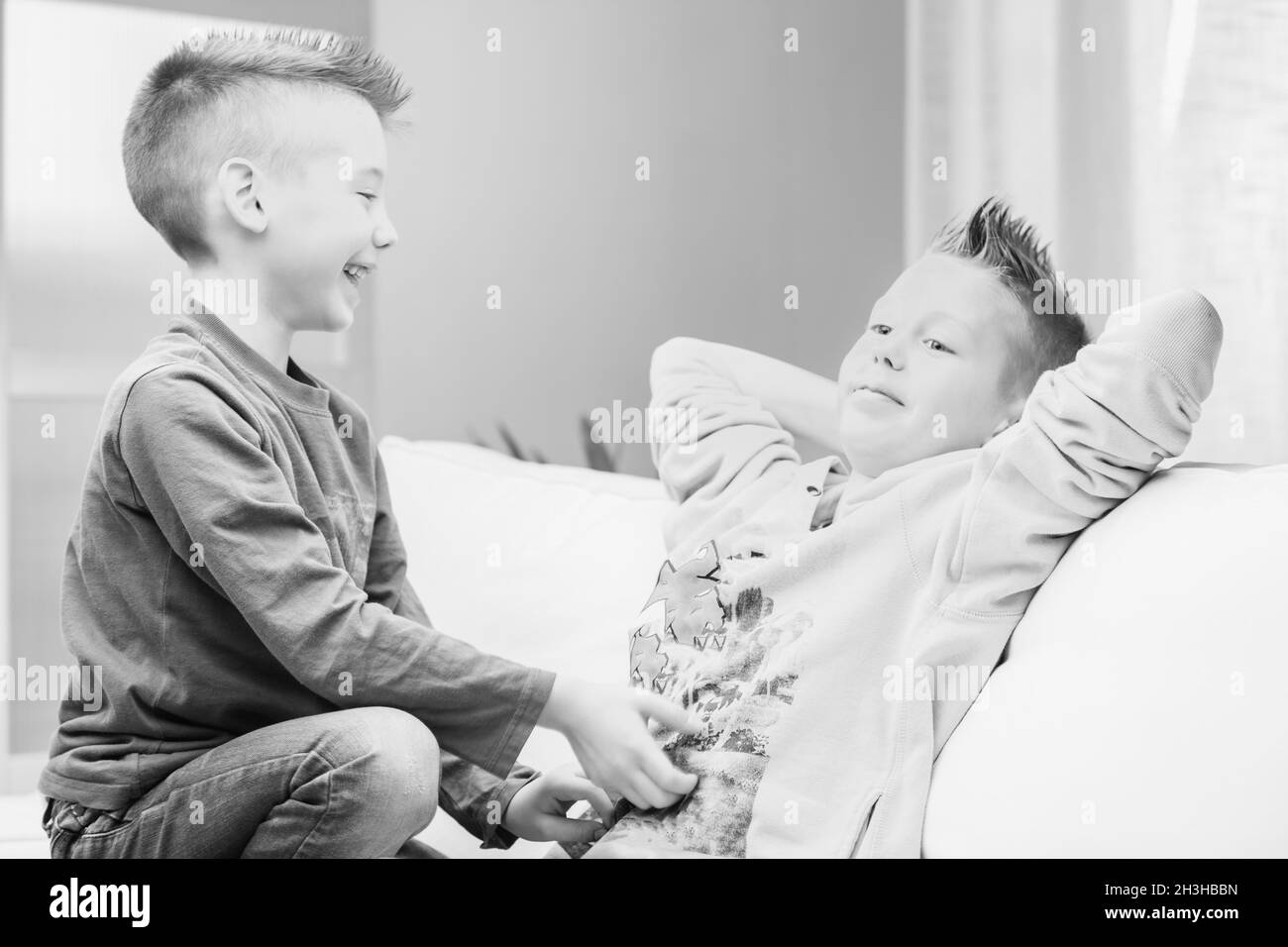 Greyscale image of two young brothers having fun playing together on a sofa at hole laughing and tickling each other Stock Photo