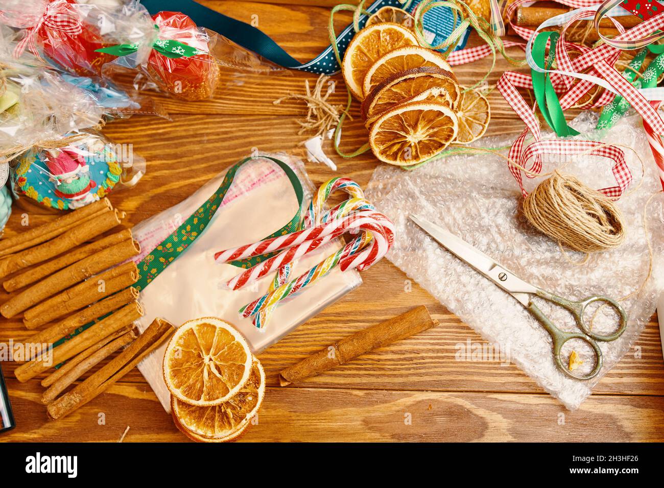 Wooden table with Christmas decor. Cinnamon sticks, candy canes, souvenirs, slices of dried oranges, colorful ribbons, scissors and wrapper. New Year's composition. Gift packaging. Stock Photo