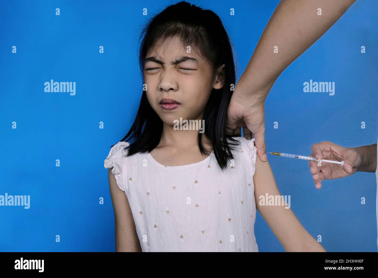A cute young Asian girl in white dress in getting a flu vaccination shot on her shoulder by a doctor. Girl feeling scare at the hospital, closing her Stock Photo
