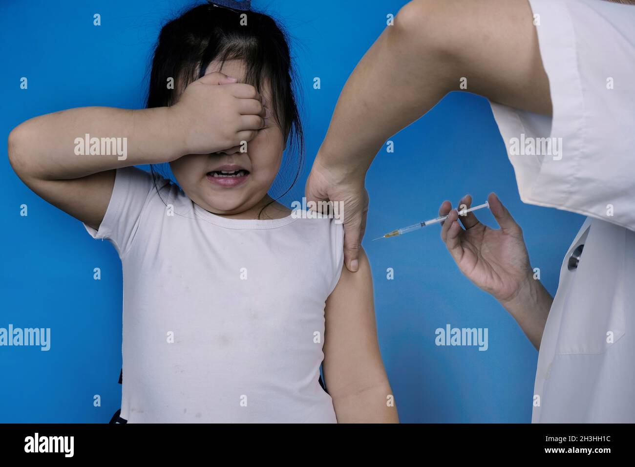 Little kid terrified by injection at the hospital. Girl afraid of syringe needle covers face while getting flu vaccine at pediatric clinic. Blue backg Stock Photo