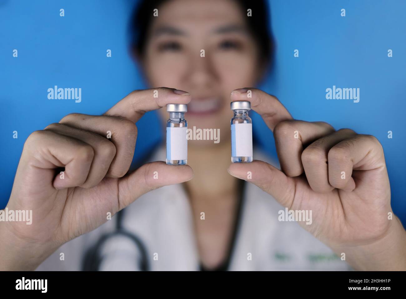A female doctor holding 2 vials of vaccine in parallel. A physician in white coat and stethoscope around her neck showing 2 glass bottles with blank w Stock Photo