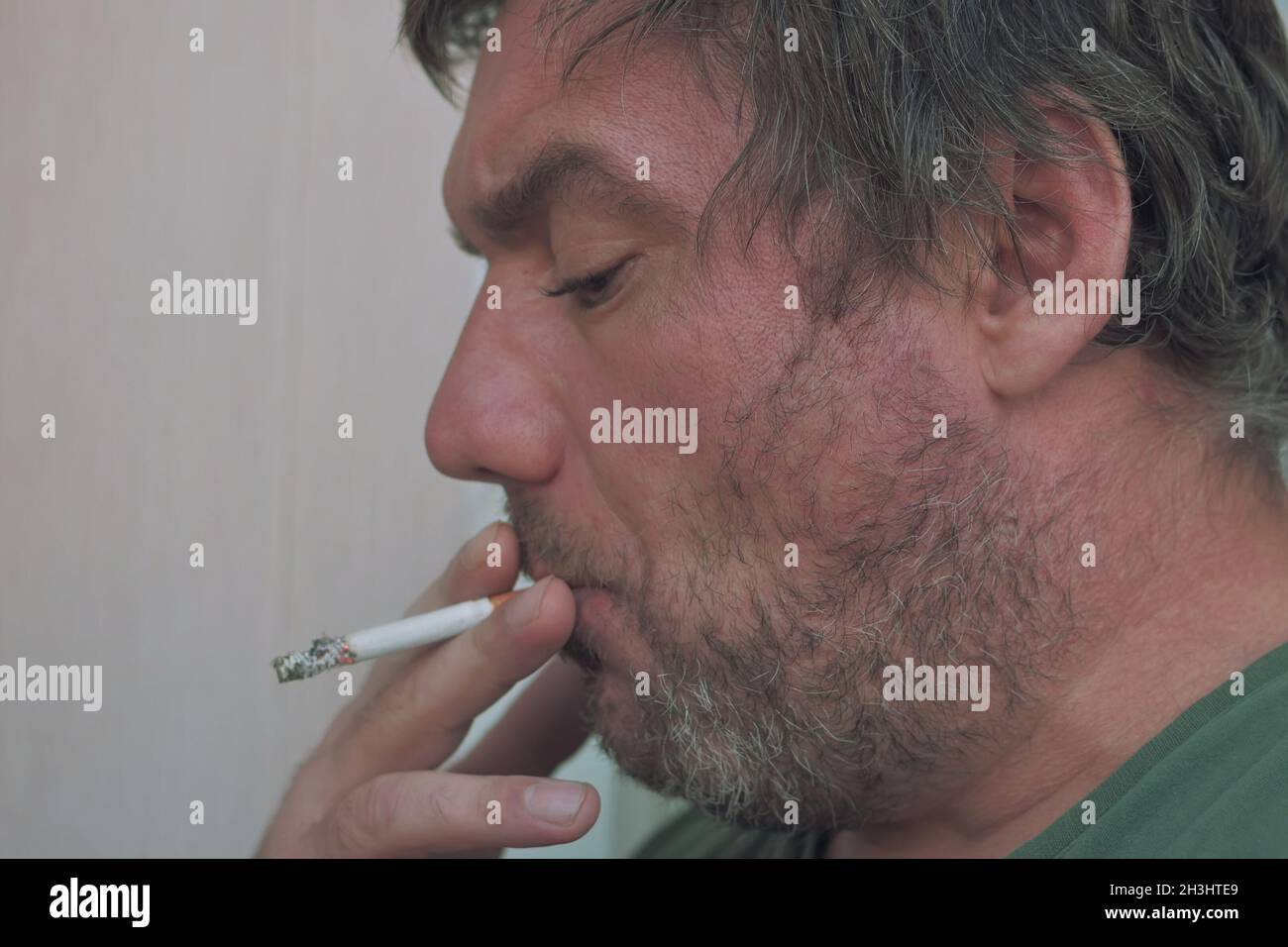 a homeless man with an old face smokes a cigarette and looks at the floor. Stock Photo