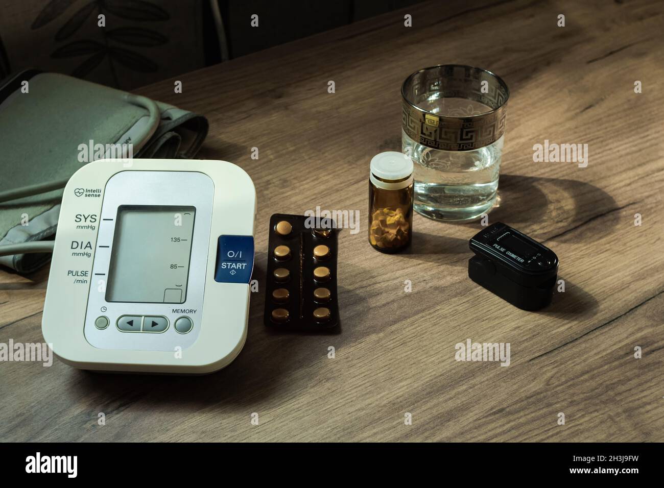 On a wooden table there is an automatic tonometer, a glass of water, pills, and an oxyometer. Health Concept Stock Photo