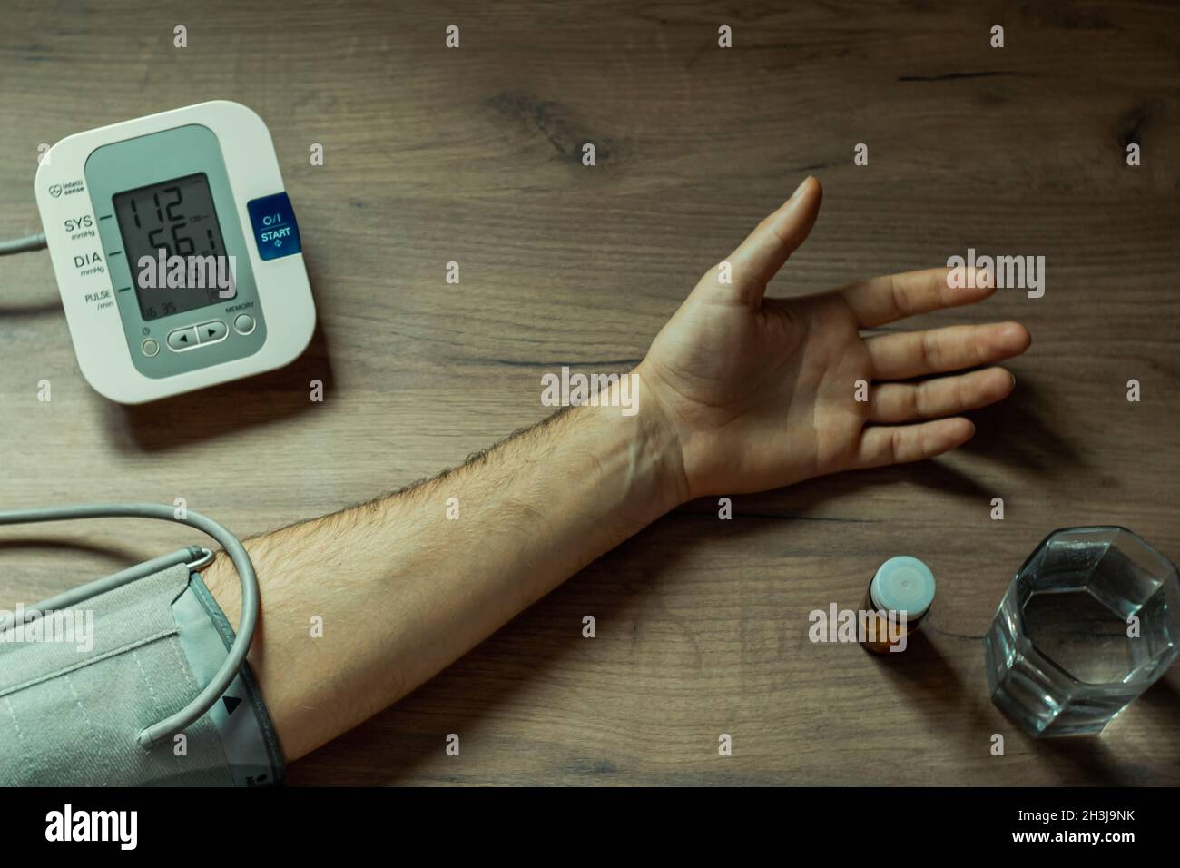 A man's hand, a glass of water, pills and a blood pressure measuring device on a wooden table. The tonometer screen shows a lowered blood pressure rea Stock Photo