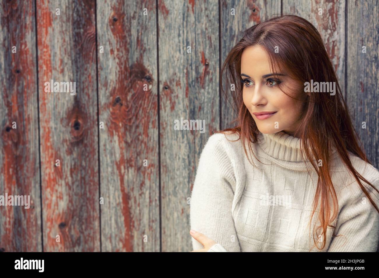 Outdoor portrait of beautiful girl or young woman with red hair wearing a white jumper leaning against an old wooden background Stock Photo