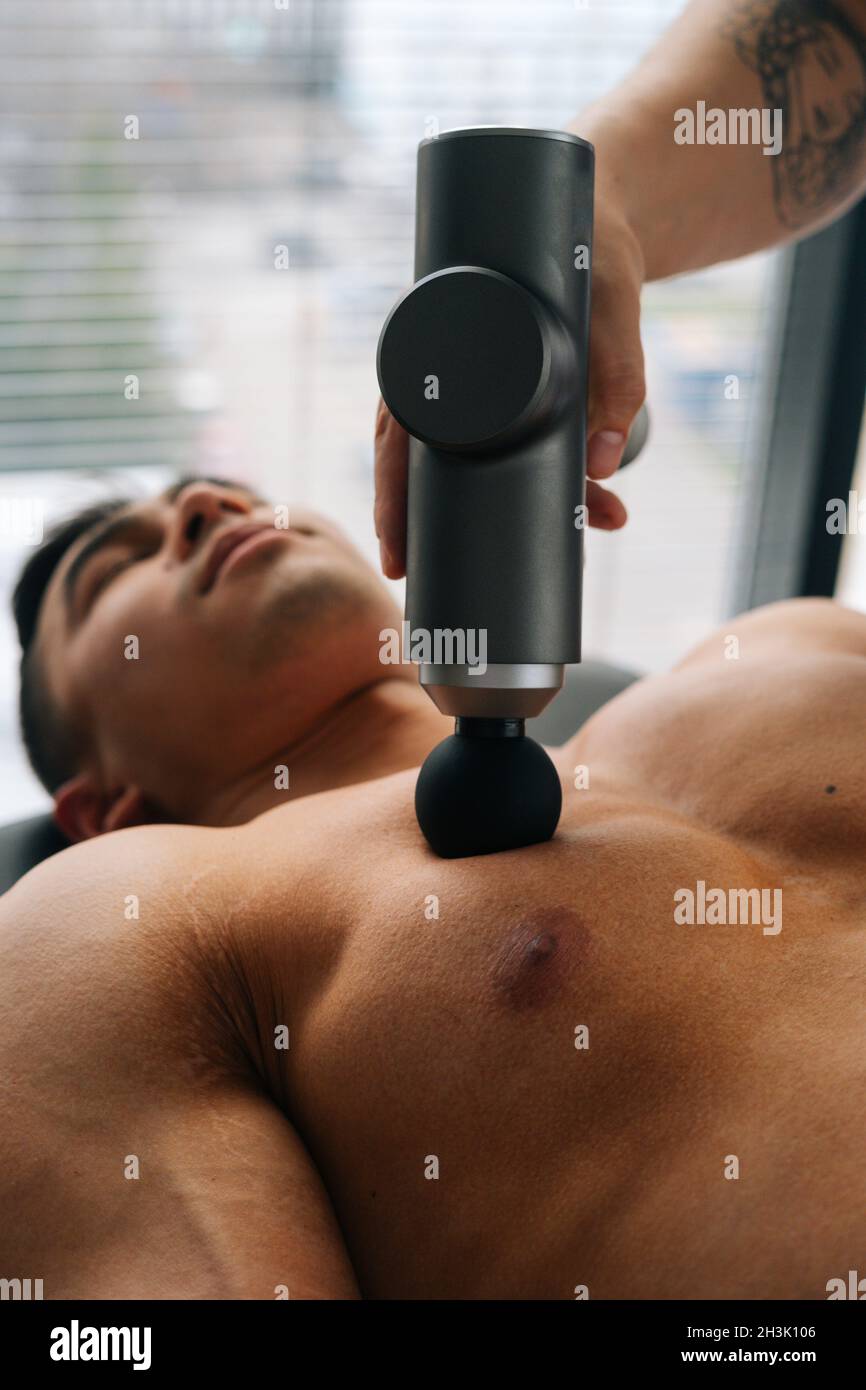 Medium shot of unrecognizable professional male masseur receiving chest muscles using massage gun percussion tool of muscular athlete man Stock Photo