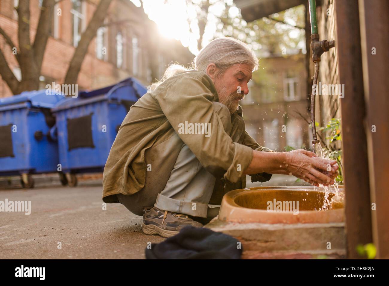 Squating old tramp cleaning face under water Stock Photo - Alamy
