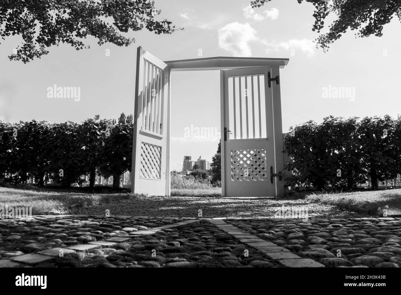 Grayscale shot of an open gate of a garden with trees under a clear sky Stock Photo