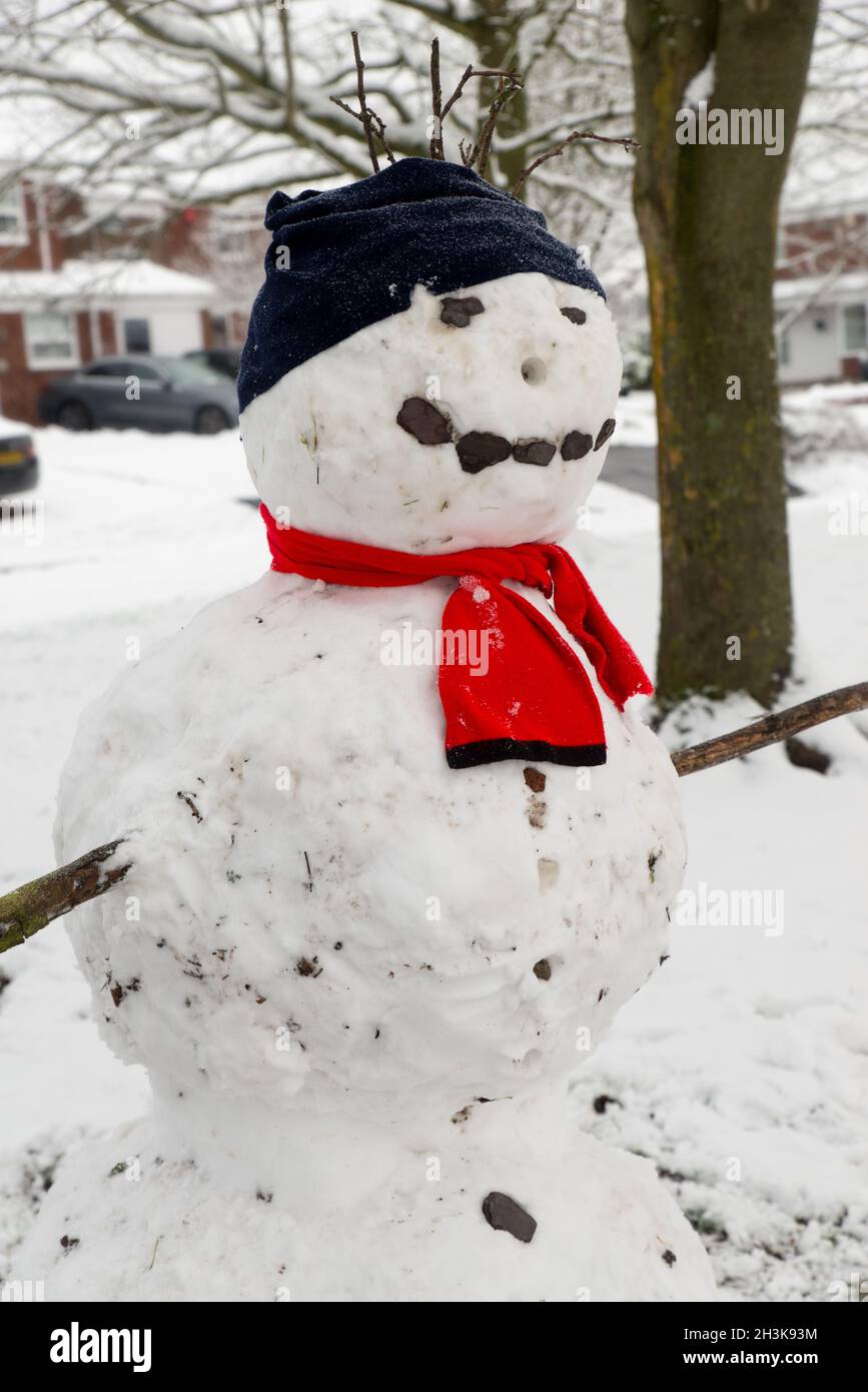 close up of large snowman built by children wearing black hat and red scarf.  two large snowballs, with twigs for arms and stones for eyes and smiley Stock Photo