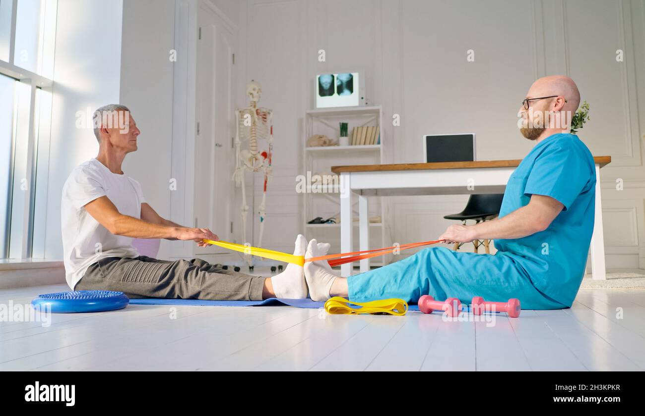 A Physical Therapist Safely Trains a Patient Using Medical Exercise Equipment at the Rehabilitation Center. Elderly Health Support Concept Stock Photo