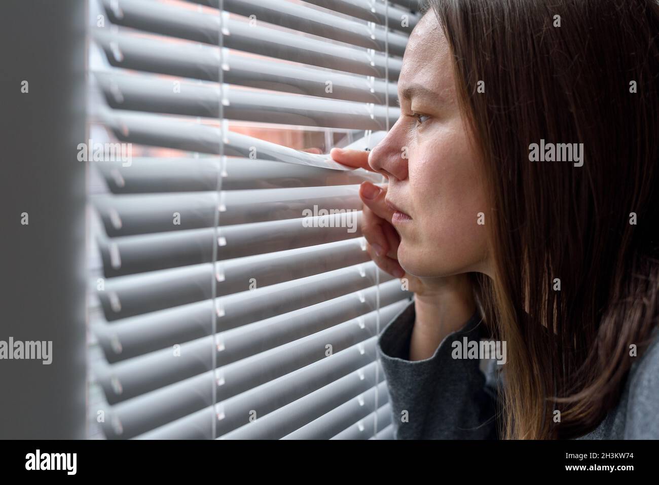 Anxious woman looking to window through blinds and waiting Stock Photo