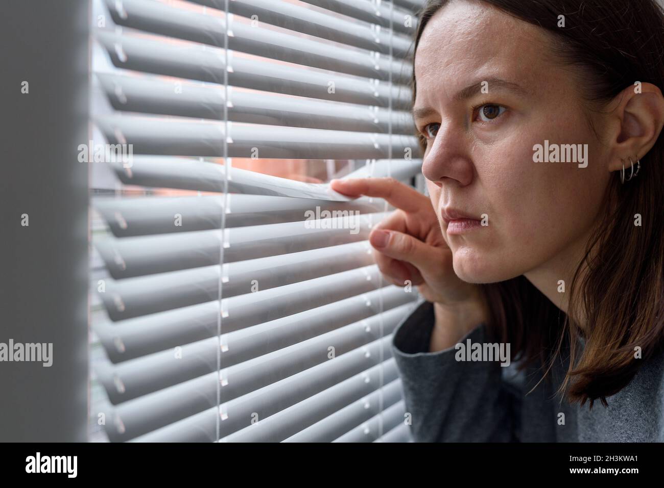 Anxious woman looking to window through blinds and waiting Stock Photo