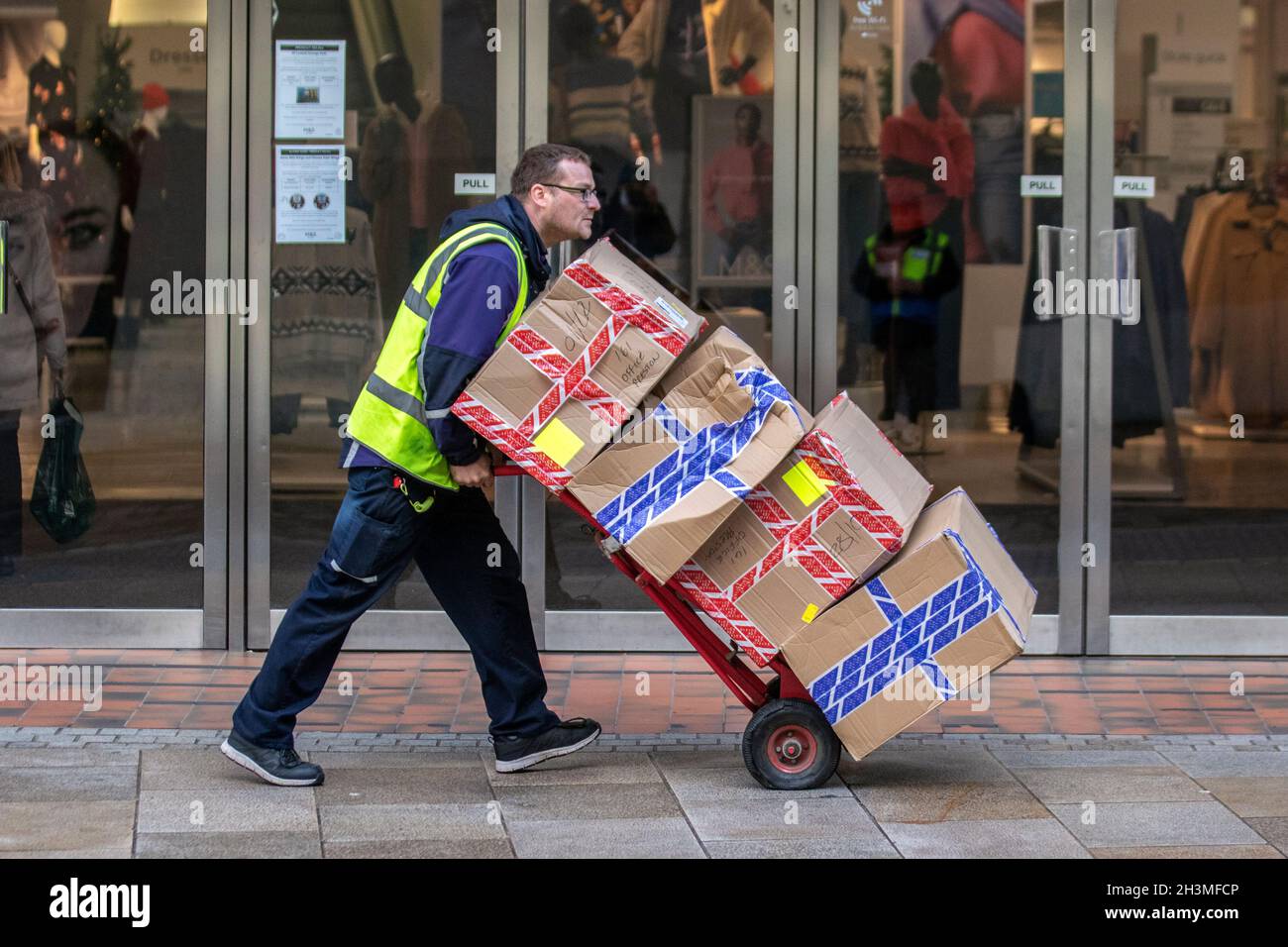 Delivery driver delivering parcels for Office store, On-trend shoe chain selling own-label and branded footwear, high street department stores sales stock on heavy duty wheeled 2 wheels sack truck. Preston, UK Stock Photo