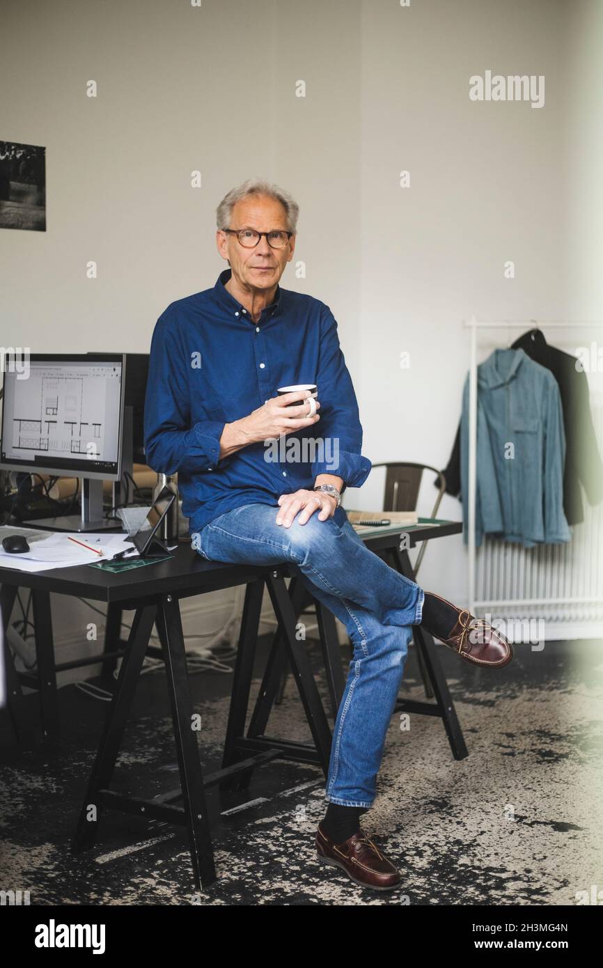Portrait of senior craftsman with coffee cup sitting on table at shop Stock Photo