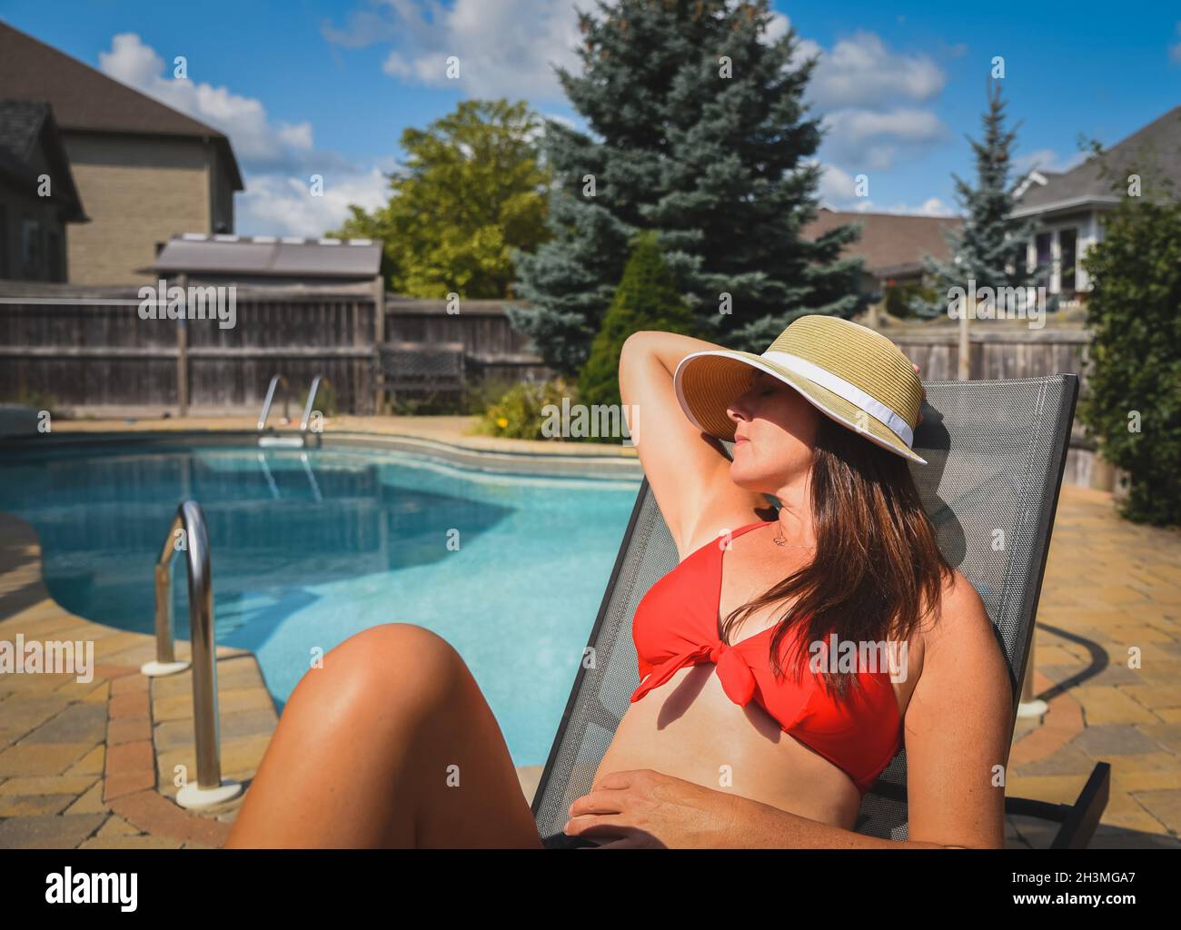 Woman in red bikini and sun hat lounging by the pool in summer. Stock Photo