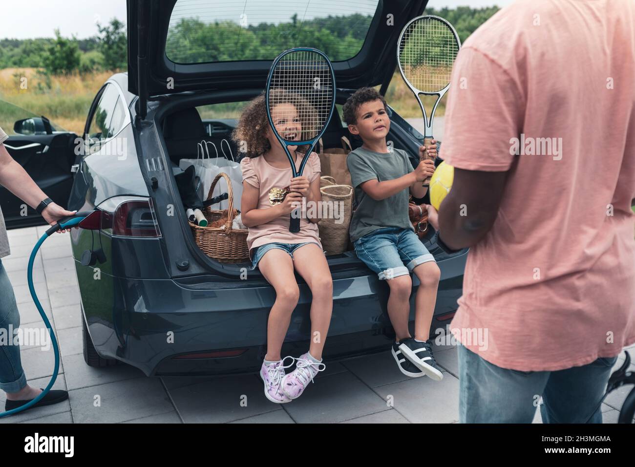 Siblings holding tennis rackets while sitting in car trunk and looking ...