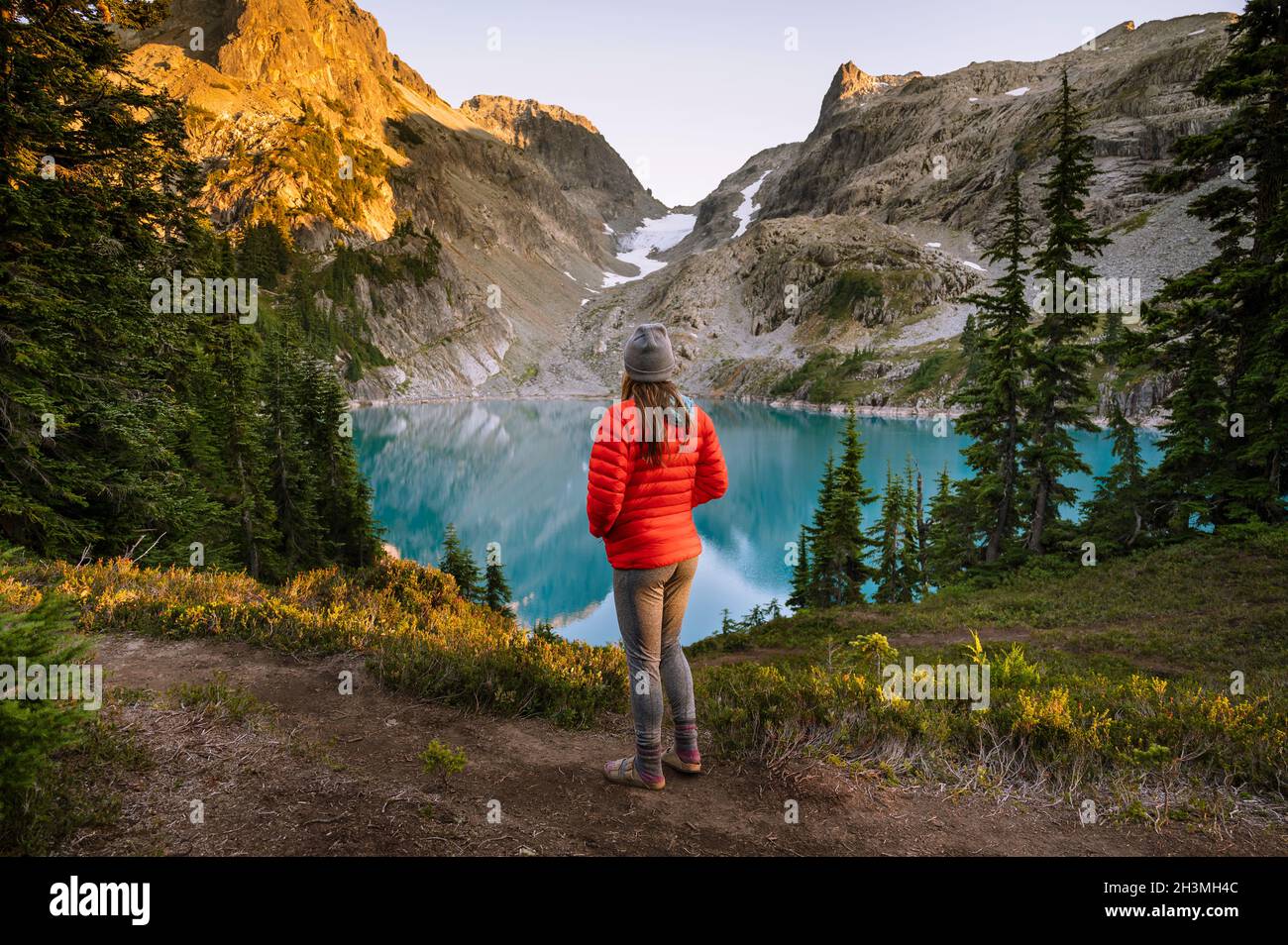 Female In Red Jacket Looking At Blue Alpine Lake At Sunset Stock Photo