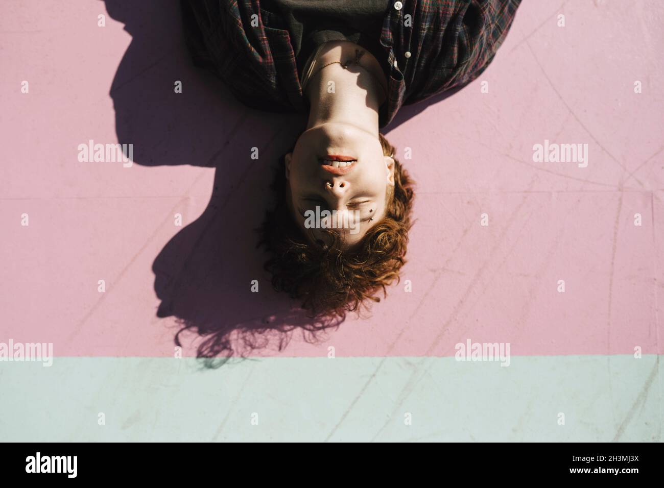 Teenage boy with eyes closed lying on pink footpath during sunny day Stock Photo