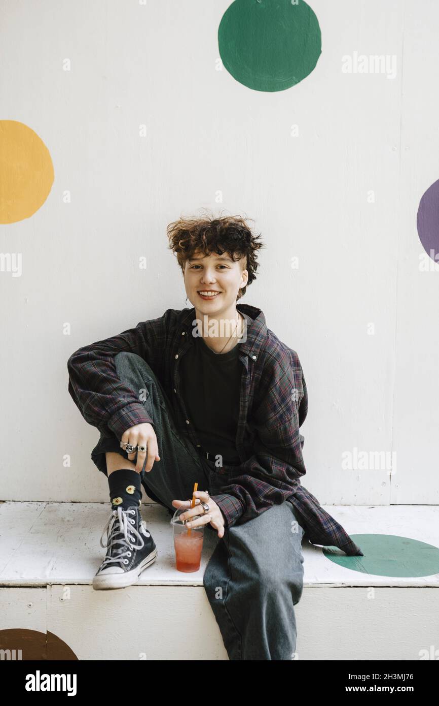 Portrait of smiling teenage boy sitting with drink on retaining wall Stock Photo