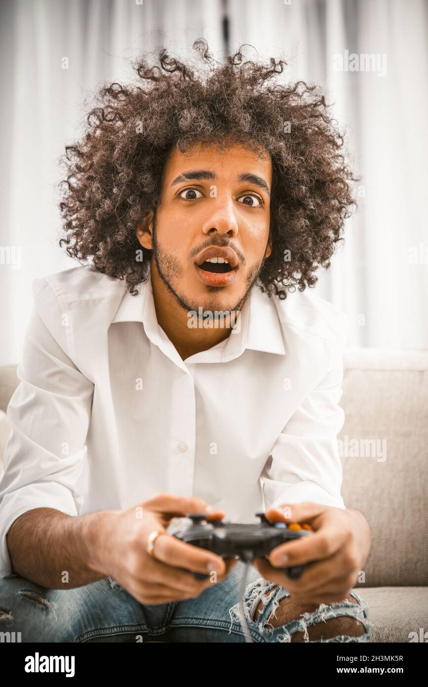 Gamer play computer game at home in self isolation. Surprised Arabic Young gay holds Play Station joystick sitting at sofa. Comp Stock Photo