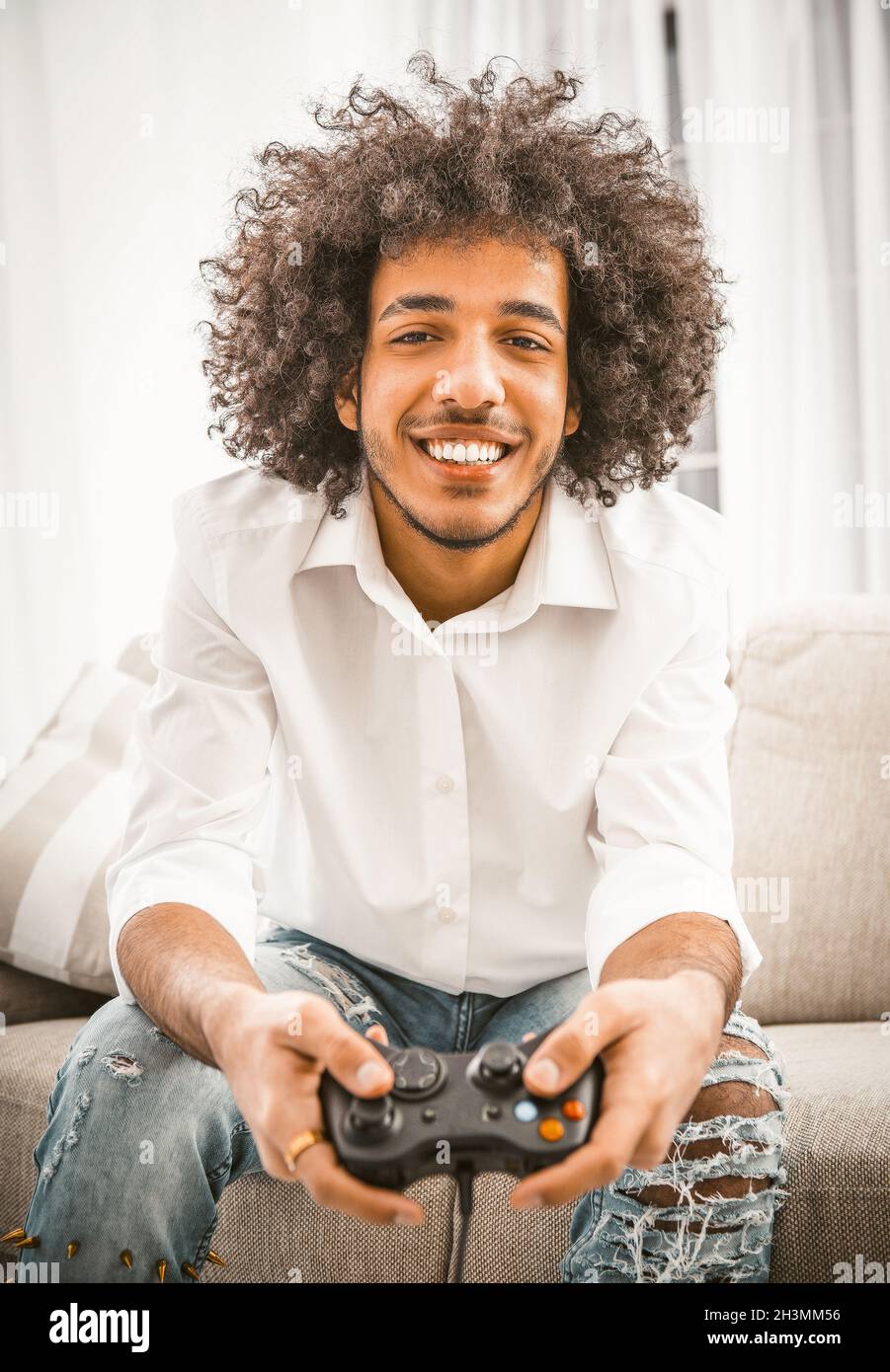 Happy Arab man smiles broadly playing computer game. Shaggy gamer holds Play Station joystick sitting at comfortable sofa at hom Stock Photo