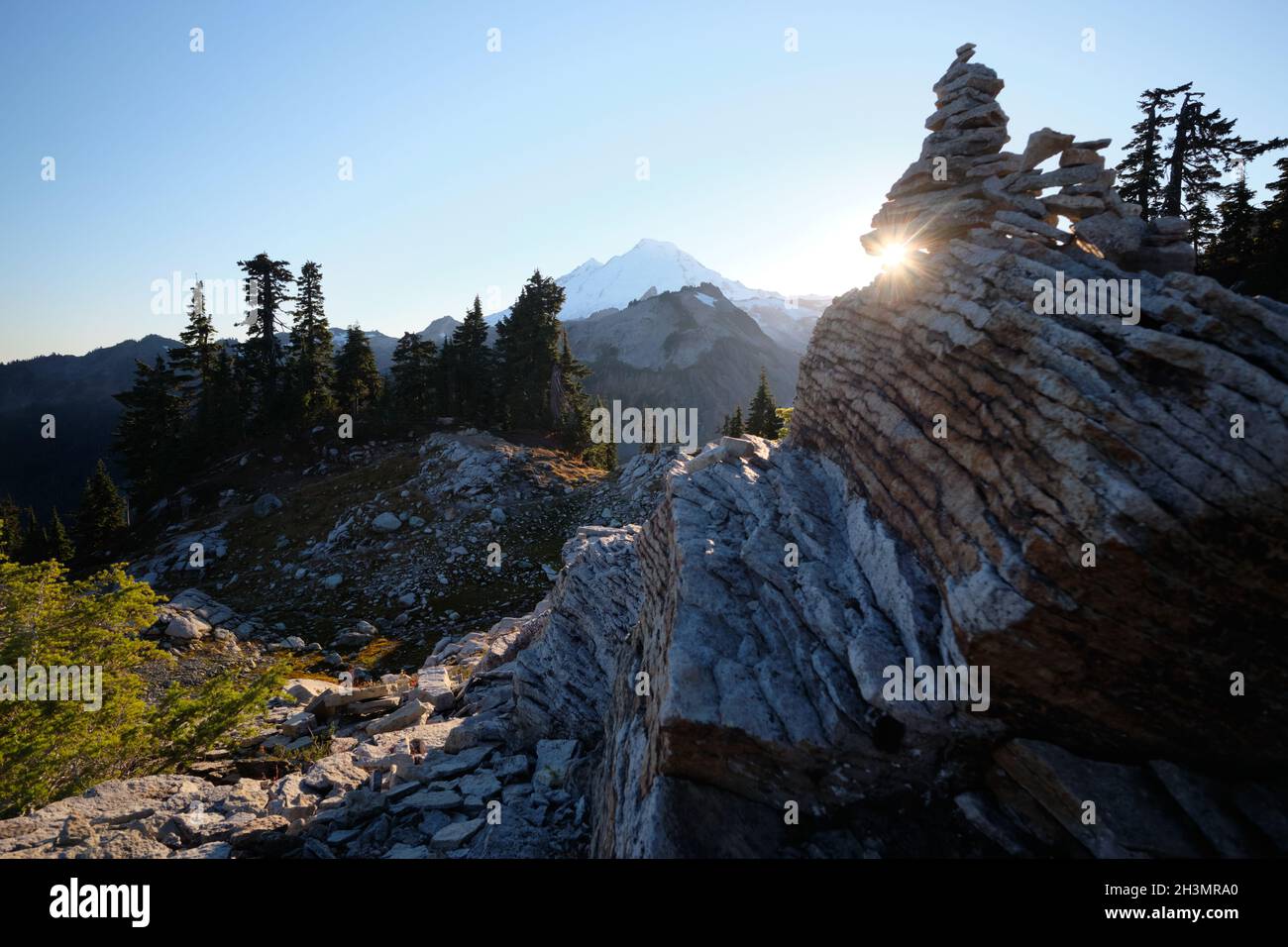 Setting sun appears as sun star through crack in layered rock on the Table Mountain Trail. Snow-covered Mount Baker, WA is on the horizon Stock Photo