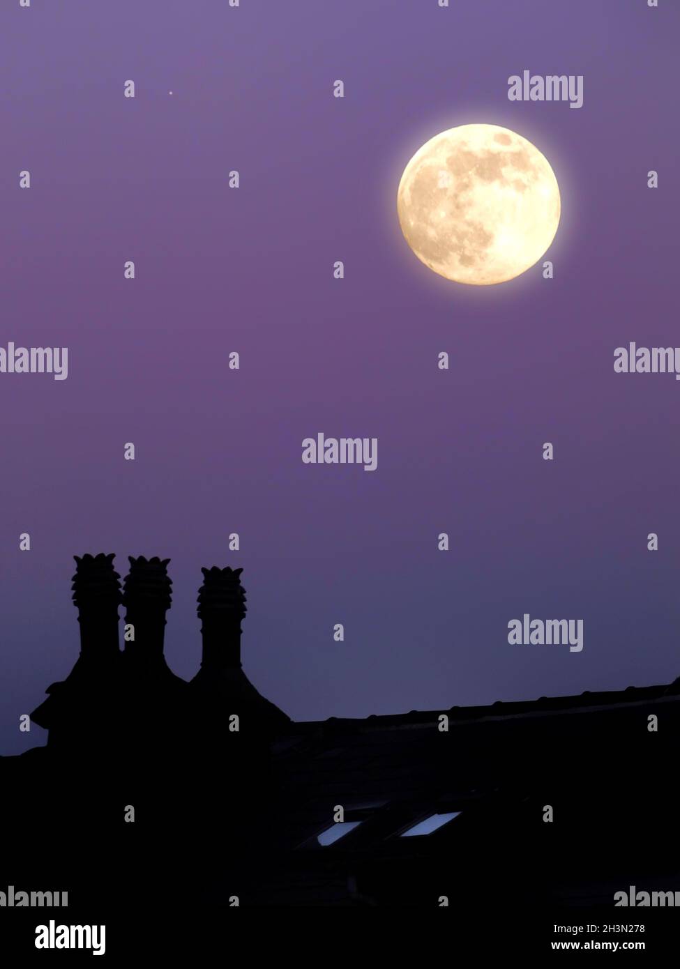 A glowing full moon in a purple twilight summer sky above a house rooftop in silhouette Stock Photo