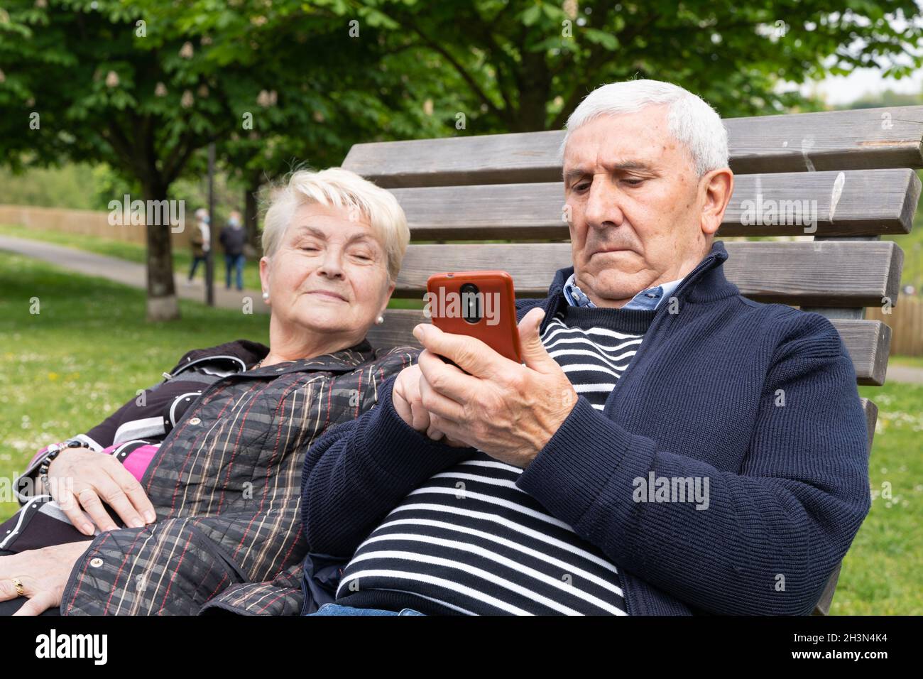 Senior couple with cellphone sitting on park bench. Elder woman peeking ...