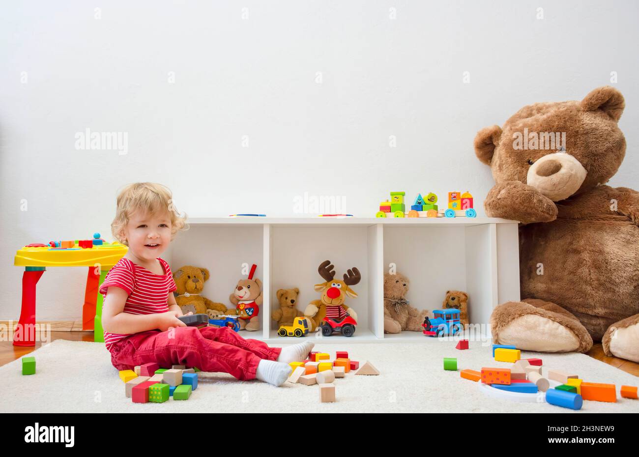 Sweet happy child boy having fun at home playing with his giant teddy bear and colorful wooden blocks and toys, on the floor. Stock Photo