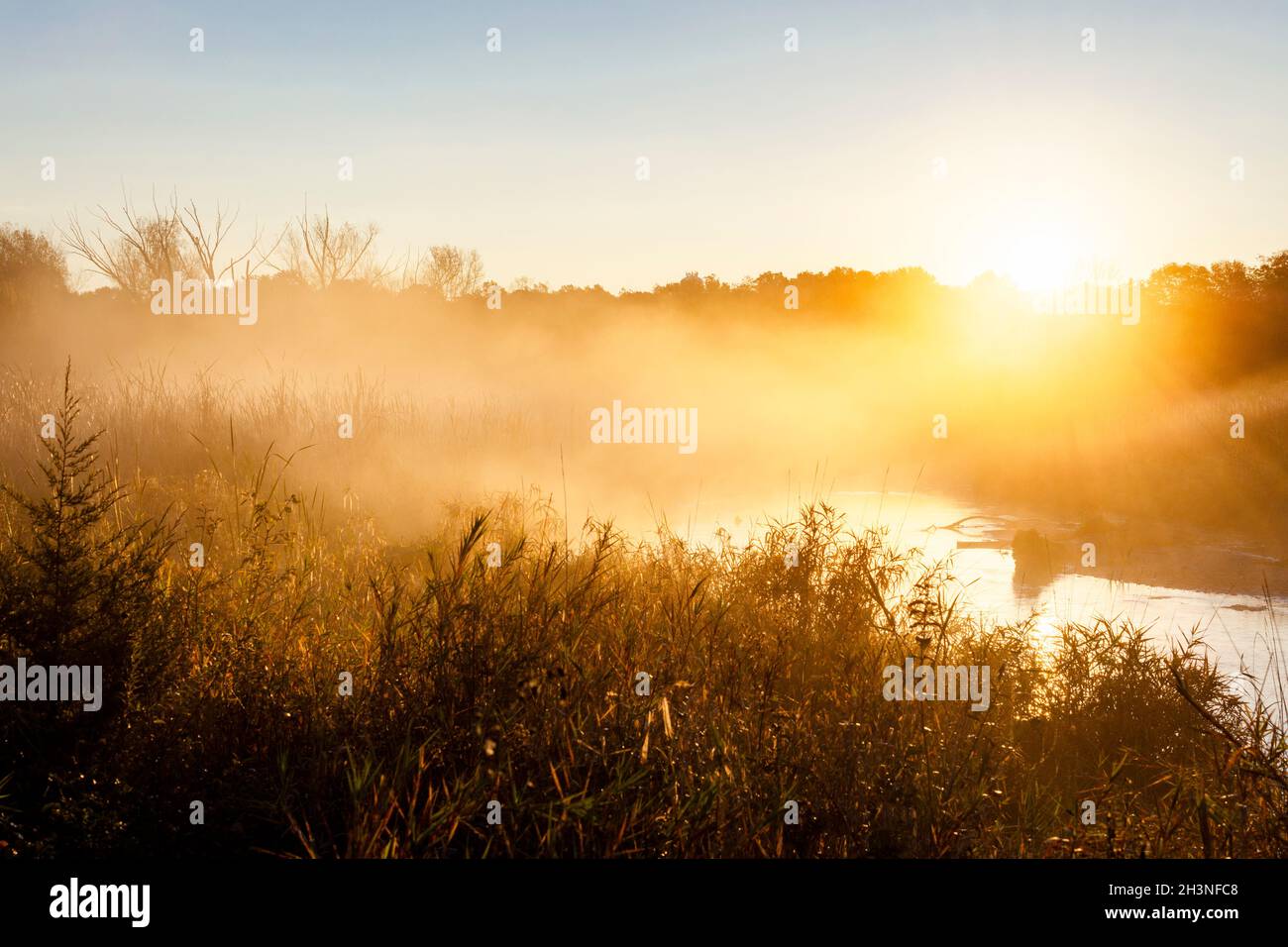 The sun peaks over the distant tree line as steam and fog collect in the marshy river basin in Waukesha County, Wisconsin on an autumn morning. Stock Photo