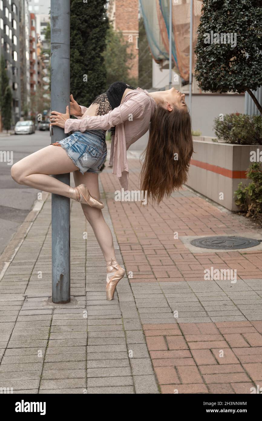 full body of a young woman leaning against a pole dancing, wearing ballet shoes, shorts and casual top, lifestyle of a ballet dancer with urban Stock Photo