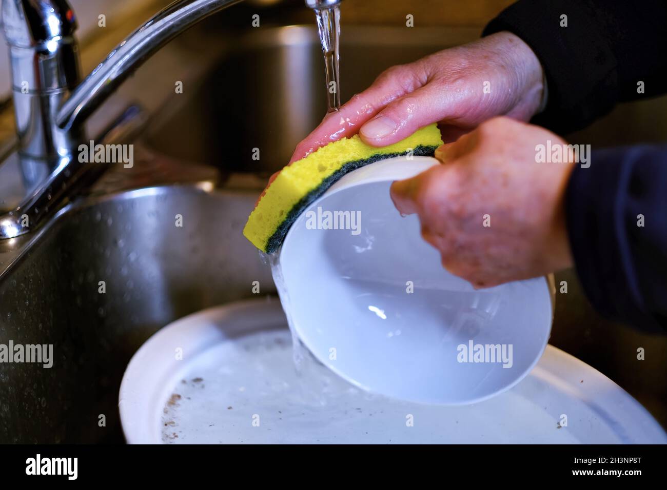 Senior man hands wash plate dishes in kitchen sink under running water at home. Authentic senior lifestyle after retirement and household work. Stock Photo