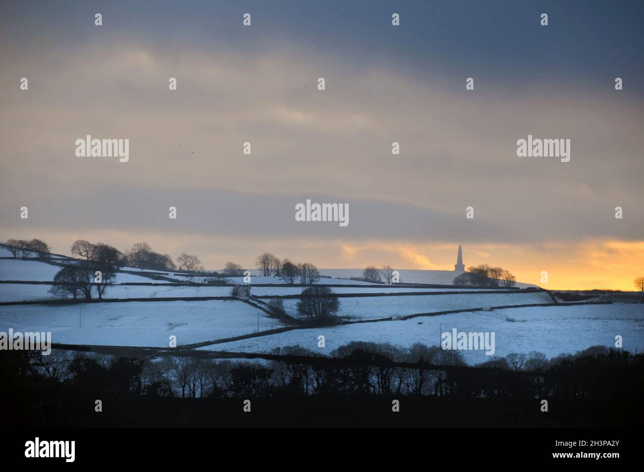Glowing orange sunset in a cloudy twilight winter sky with snow covered fields with stoodley pike monument in the distance in ca Stock Photo